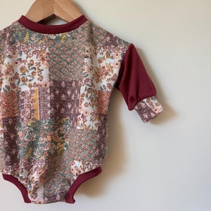 以下が含まれることがあります： クリーム、茶色、オレンジの色合いの、花柄と幾何学模様のパッチワークデザインのベビーボディスーツ。長袖とトリムは濃いバーガンディ色です。ボディスーツは木製のハンガーに吊るされています。