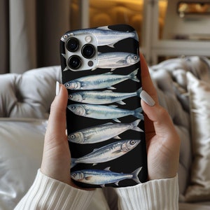 以下が含まれることがあります： 黒い電話ケースに、黒い背景に銀色の魚のデザインが施されています。電話ケースにはカメラレンズの切り抜きがあります。