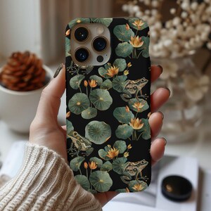 May include: A black phone case with a floral and frog design. The case has a green, brown, and gold color scheme. The case has a camera lens cutout and a cutout for the phone's buttons.