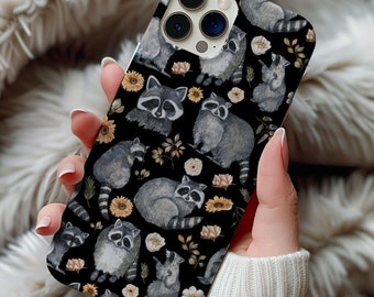 以下が含まれることがあります： 黒いスマートフォンケースで、グレーと白のアライグマが黄色いヒマワリや小さなベージュの花と一緒に繰り返されるパターンが描かれています。ケースの左上には、3つのレンズを備えたカメラシステム用の切り抜きがあります。
