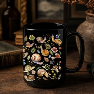 May include: A black ceramic mug featuring a repeating pattern of various snails, small mushrooms, green leaves, tiny blue berries, and small yellow stars scattered across the surface. The mug has a curved handle and a smooth, glossy finish.