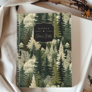 May include: A hardcover notebook with a forest scene cover. The cover features a pattern of green and white trees with the text "Notebook" and "Clara's Notes" in a black frame. The notebook is on top of a white book.