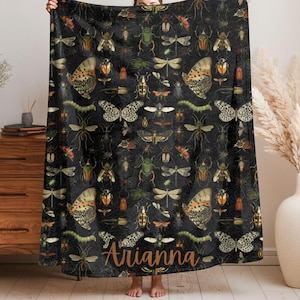 May include: Black blanket featuring a detailed pattern of various insects, including butterflies, beetles, and dragonflies in shades of green, orange, and cream. The blanket has the name "Arianna" in a brown script font at the bottom.