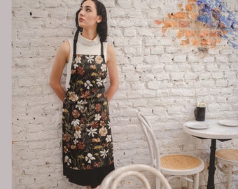 May include: A woman wearing a black and white floral apron with a white turtleneck shirt. The apron has a black ruffle at the bottom. The woman is standing in front of a white brick wall with a white table and chairs in the background.