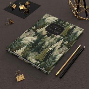 May include: A hardcover notebook with a forest pattern in shades of green, white, and beige. The cover features the text "Notebook" and "Chris's Notes" in a decorative frame. Gold and black pencils and gold binder clips are also visible.