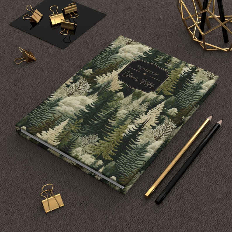 May include: A hardcover notebook with a forest pattern in shades of green, white, and beige. The cover features the text "Notebook" and "Chris's Notes" in a decorative frame. Gold and black pencils and gold binder clips are also visible.
