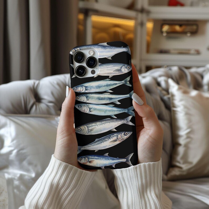 以下が含まれることがあります： 黒い電話ケースに、黒い背景に銀色の魚のデザインが施されています。電話ケースにはカメラレンズの切り抜きがあります。