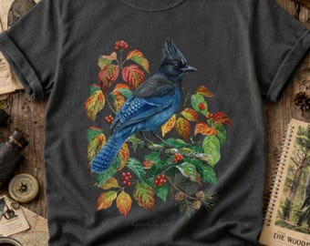 May include: A charcoal gray short-sleeve t-shirt featuring a detailed illustration of a blue Steller's jay perched on a branch. The bird is surrounded by vibrant autumn-colored leaves in shades of red, orange, and green, along with small red berries.