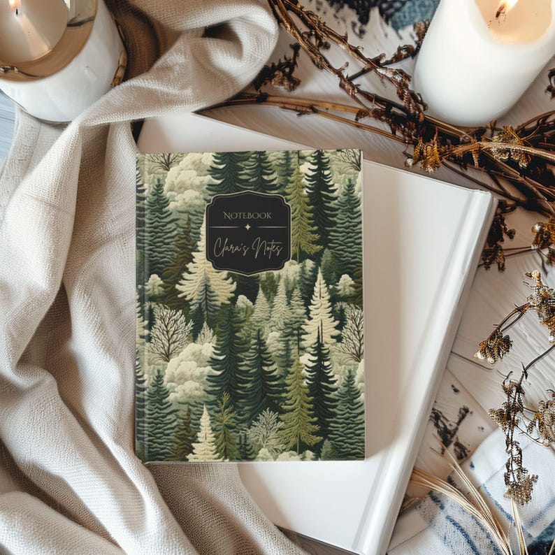May include: A hardcover notebook with a forest scene cover. The cover features a pattern of green and white trees with the text "Notebook" and "Clara's Notes" in a black frame. The notebook is on top of a white book.