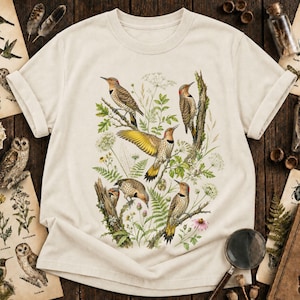 May include: A cream-colored short-sleeve t-shirt featuring a detailed botanical and wildlife illustration. The graphic displays five northern flicker birds perched on branches and flying amidst green foliage, ferns, and white wildflowers.