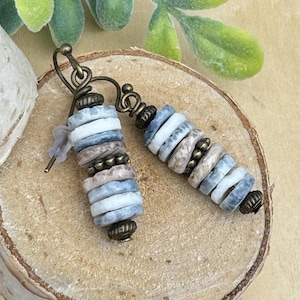 May include: A pair of handcrafted earrings featuring stacked, round beads in shades of white, blue, and tan. The beads are strung together with small, bronze-colored metal beads and end caps. The earrings have a bronze-colored hook.