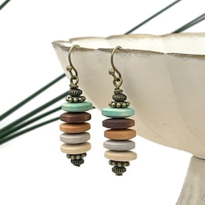 May include: A pair of dangle earrings with a stack of five wooden beads in shades of brown, beige, and teal. The beads are separated by small bronze beads and the earrings have bronze hooks.