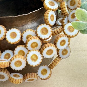 May include: A collection of small, round, daisy-shaped beads with a white center and a textured, light brown outer edge. The beads are scattered near a bronze-colored bowl and a sprig of green leaves.