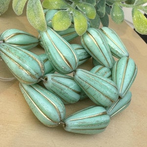 May include: A collection of teal green glass beads with gold accents. The beads are shaped like teardrops and have a textured surface.