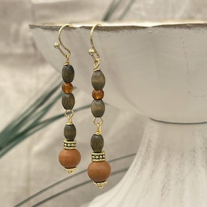 May include: A pair of long, dangle earrings with brown and green wooden beads and gold accents. The earrings have a brown wooden bead at the bottom.