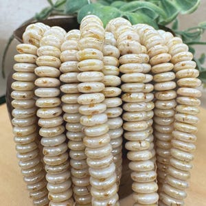 May include: Strands of off-white, disc-shaped beads. The beads have a slightly irregular shape and a glossy finish, with subtle variations in color. The beads are strung together, creating a flowing, textured appearance. The background includes a blurred plant and a brown pot.