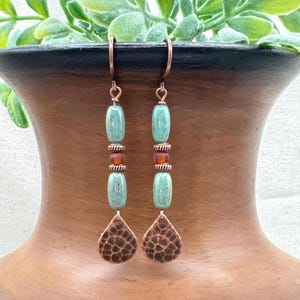 May include: A pair of handcrafted earrings with copper-colored ear wires and teardrop-shaped pendants. The earrings feature turquoise-colored beads, amber-colored beads, and textured copper pendants. The earrings are designed for pierced ears.