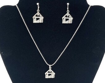 May include: A silver necklace and earrings set featuring a nativity scene pendant and matching earrings. The pendant and earrings are shaped like a small house with a star on top and a scene of the nativity inside.