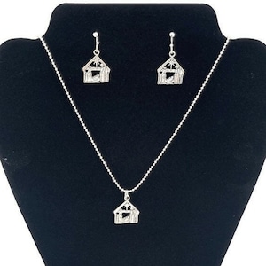 May include: A silver necklace and earrings set featuring a nativity scene pendant and matching earrings. The pendant and earrings are shaped like a small house with a star on top and a scene of the nativity inside.