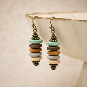 May include: A pair of dangle earrings featuring stacked circular beads in turquoise, dark brown, tan, gray, and cream colors. The beads are accented with small, textured bronze-toned metal spacers and hang from curved bronze-toned metal ear wires.