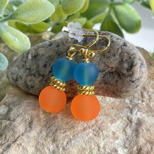 May include: A pair of dangle earrings with two beads on each earring. The top bead is a light blue color and the bottom bead is a bright orange color. The beads are frosted glass and the earring hooks are gold.