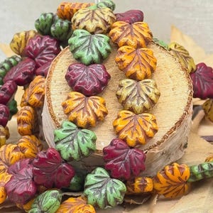 May include: Close-up of leaf-shaped beads in various colors, including green, yellow, orange, and burgundy. The beads are arranged on a wooden surface, showcasing their intricate details and autumnal theme. These beads are ideal for jewelry making.
