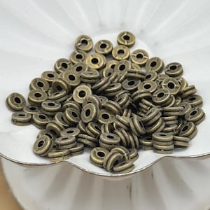 May include: A pile of antique brass-colored metal beads with a circular shape and a central hole. The beads have a ridged design around the edges. The beads are scattered on a white, scalloped-edge dish.