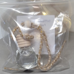 May include: A clear glass diffuser bottle with a wooden cap and a braided rope, packaged in a clear plastic bag. The rope is beige with gold accents. A white label with text is visible inside the bag.