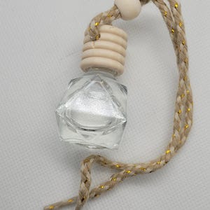 May include: A small, clear glass bottle with a faceted design and a silver interior. It has a wooden cap and a braided beige cord with gold threads. The bottle is likely for fragrance or essential oils and is suspended by the cord.
