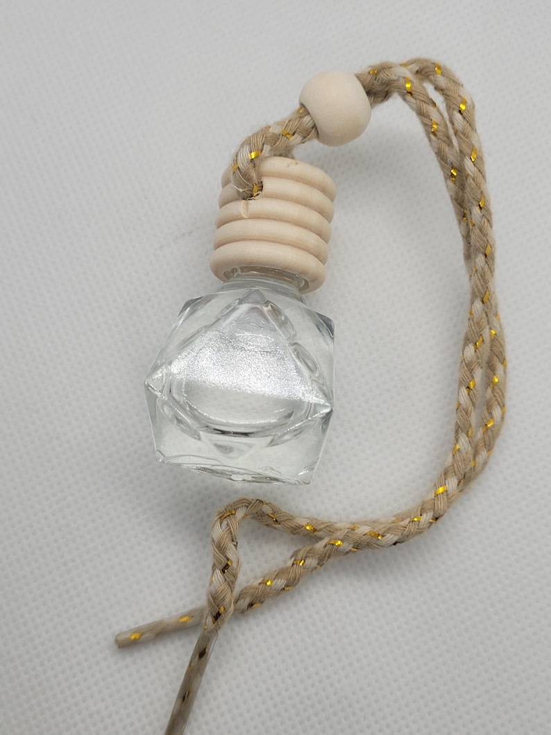 May include: A small, clear glass bottle with a faceted design and a silver interior. It has a wooden cap and a braided beige cord with gold threads. The bottle is likely for fragrance or essential oils and is suspended by the cord.