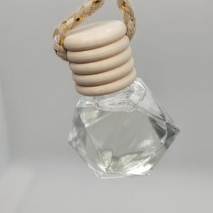 May include: A clear, faceted glass bottle with a wooden cap and a braided rope hanger. The wooden cap is made of stacked, circular pieces. The rope has gold accents. The bottle is empty, suggesting it is for essential oils or perfume.