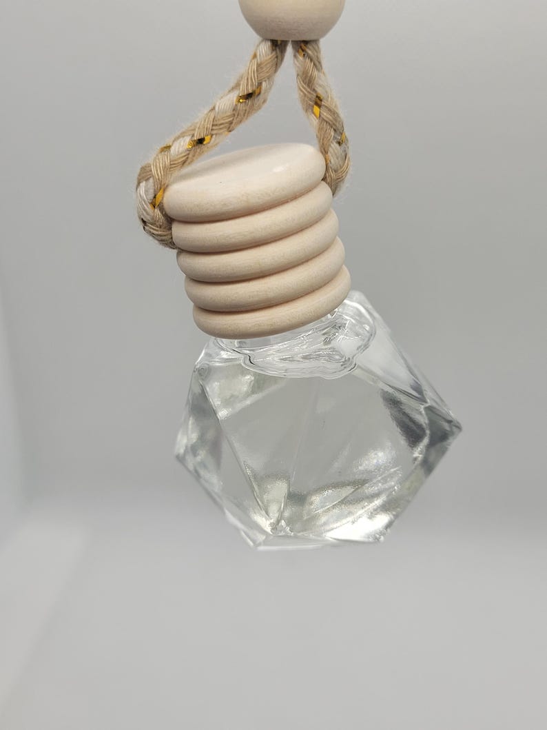 May include: A clear, faceted glass bottle with a wooden cap and a braided rope hanger. The wooden cap is made of stacked, circular pieces. The rope has gold accents. The bottle is empty, suggesting it is for essential oils or perfume.