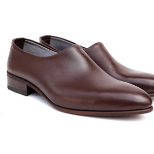 May include: A pair of brown leather slip-on shoes. The shoes have a low heel and a rounded toe. The leather has a polished finish. The shoes are set against a white background.