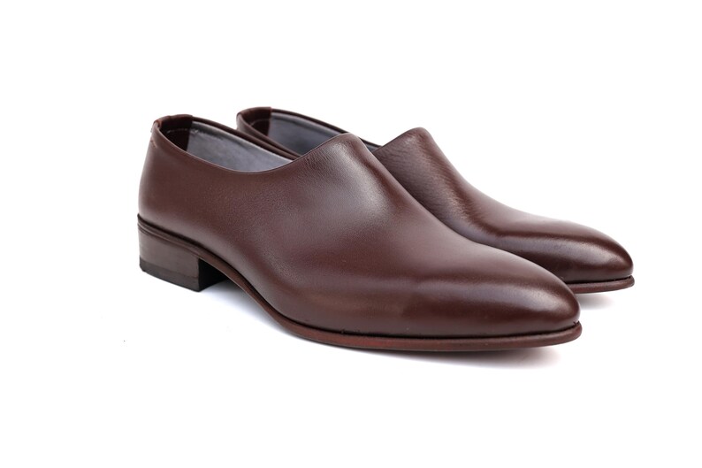 May include: A pair of brown leather slip-on shoes. The shoes have a low heel and a rounded toe. The leather has a polished finish. The shoes are set against a white background.