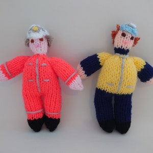 Pode incluir: Duas bonecas de malha vestindo roupas de cores diferentes. Uma boneca está vestindo um macacão vermelho com um chapéu branco e a outra boneca está vestindo uma jaqueta amarela com calças azuis e um chapéu azul.