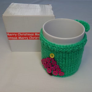 Op de afbeelding: Een witte keramische mok met een groene gebreide cozy met een roze en gele knoop kerstboom ontwerp. De cozy heeft de tekst "Merry Christmas" erop.