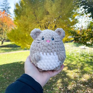 May include: A crocheted plush toy shaped like a mouse with a white belly and gray fur. The toy has black eyes, a pink nose, and a black stitched mouth.