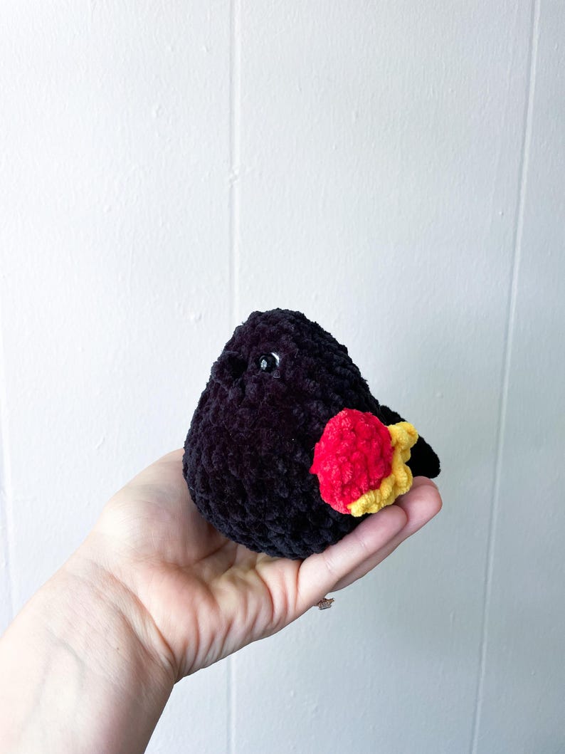 May include: A black crocheted bird with a red and yellow wing. The bird is small and has a fluffy texture.