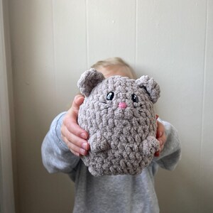 May include: A gray crocheted plush toy shaped like a mouse with pink embroidered eyes and nose. The toy is being held by a child.