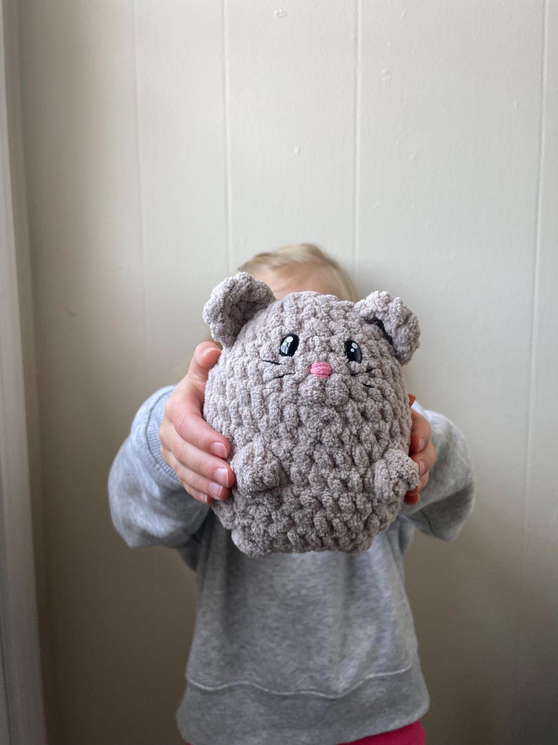 May include: A gray crocheted plush toy shaped like a mouse with pink embroidered eyes and nose. The toy is being held by a child.