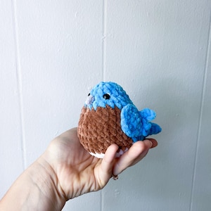 May include: A small crocheted bluebird with brown and white accents. The bird is sitting in a hand.