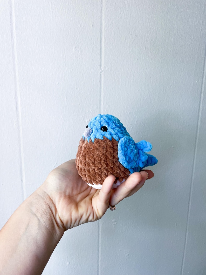 May include: A small crocheted bluebird with brown and white accents. The bird is sitting in a hand.