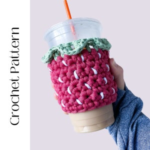 Puede incluir: Un cubrevasos de ganchillo en forma de fresa de color rosa y blanco con hojas verdes. El cubrevasos está en un vaso de plástico transparente con una pajita.