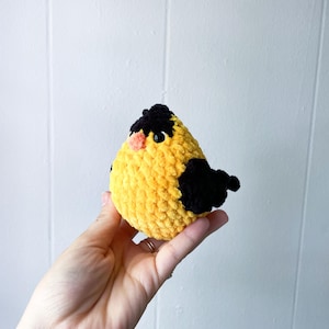 May include: A crocheted yellow bird with black accents and a pink beak. The bird is held in a person's hand.