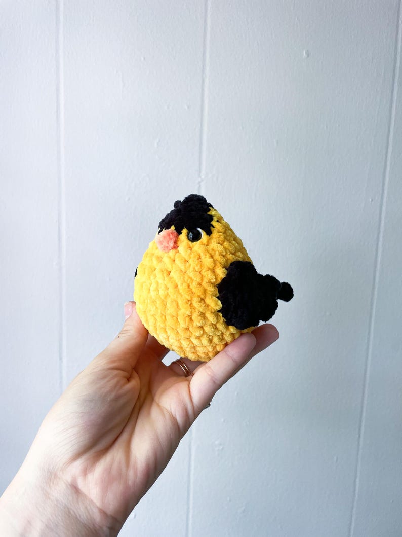 May include: A crocheted yellow bird with black accents and a pink beak. The bird is held in a person's hand.