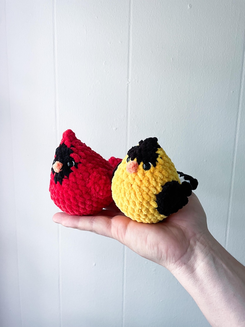 May include: Two crocheted plush birds, one red and one yellow with black accents. The birds are sitting on a hand.