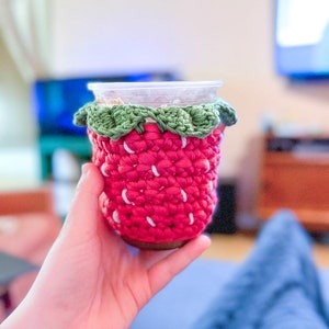 May include: A crocheted strawberry-shaped cup cozy in pink and green. The cozy is made of yarn and has a white stripe pattern.