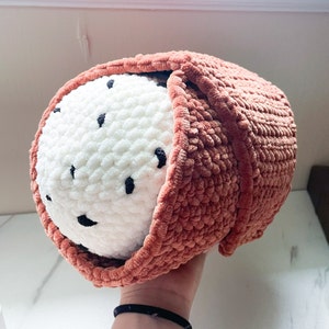 May include: A crocheted plush toy shaped like a sushi roll. The toy is made of a soft, pink yarn and has a white crocheted filling. The toy has black details that resemble sesame seeds.