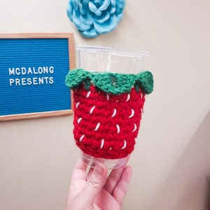 May include: A red and green crocheted strawberry-shaped cup cozy. The cozy is made of yarn and has a white stitch detail. The cozy is on a clear plastic cup.