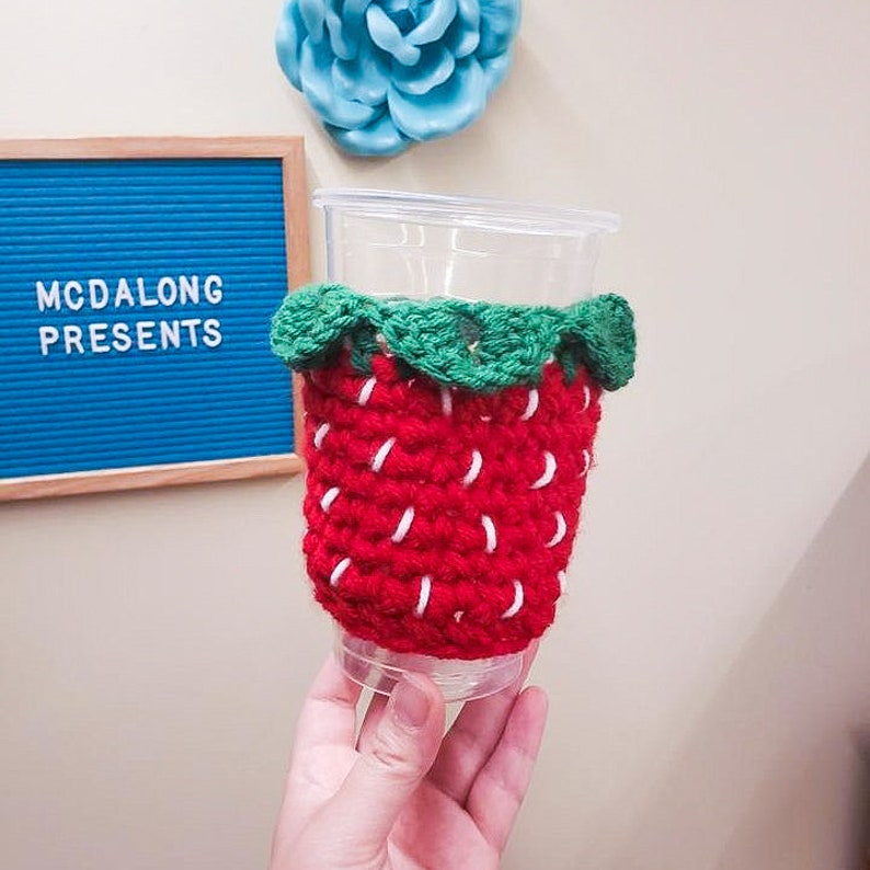 May include: A red and green crocheted strawberry-shaped cup cozy. The cozy is made of yarn and has a white stitch detail. The cozy is on a clear plastic cup.